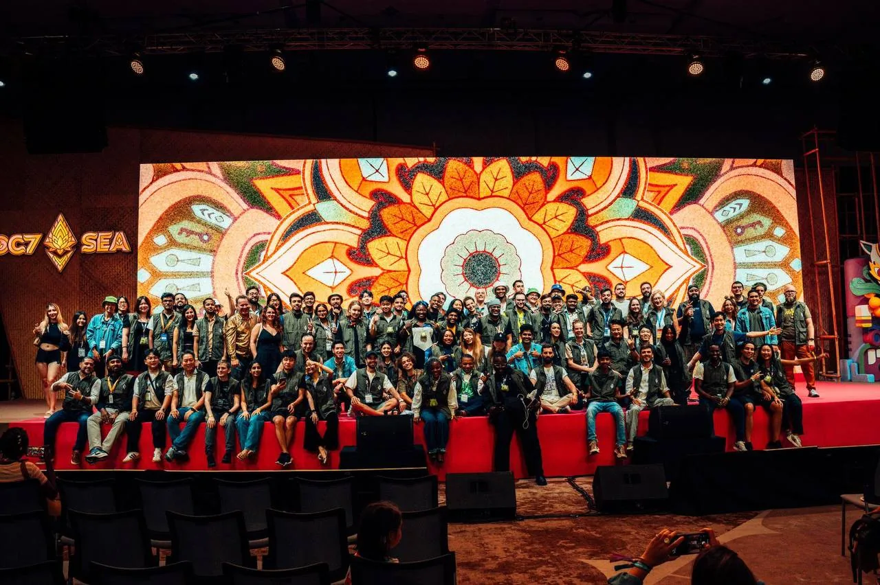 A large group of Devcon 7 SEA volunteers in olive vests posing together on a red stage in front of a glowing orange mandala screen.