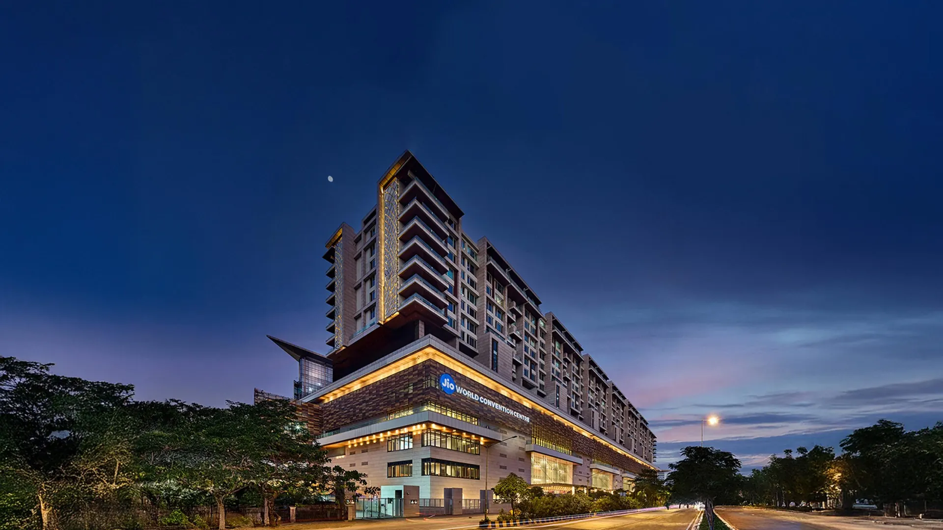 The Jio World Convention Centre in Bandra Kurla Complex at dusk, its facade lit in gold under a deep blue sky.
